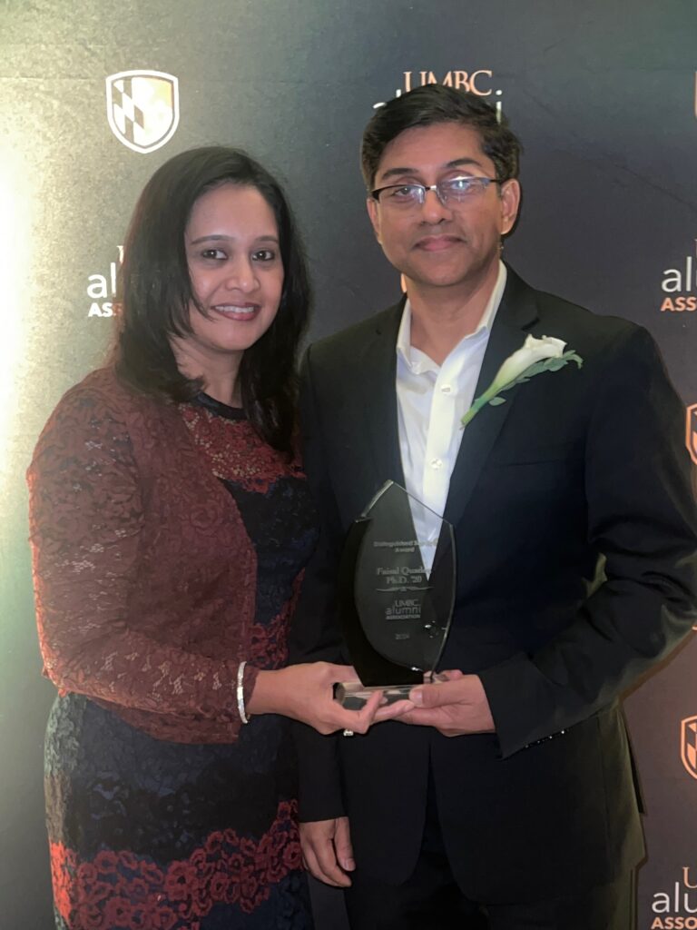 A woman and a man posing for a picture in front of a black backdrop that has the words "UMBC alumni association" written in gold and white font. The man is holding a glass award that has his name, Faisal Quader, PhD, '20. 