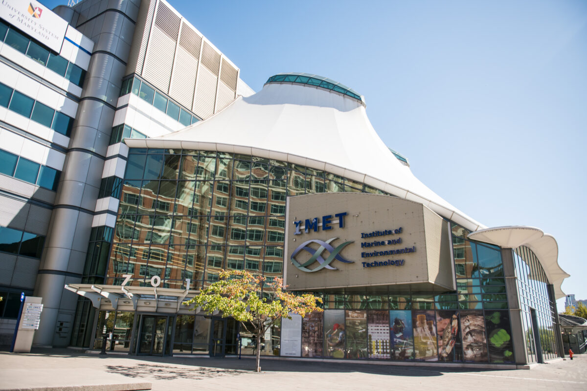 Large building with a swirly fish-shaped logo and the words "IMET Institute of Environmental Technology" on the side. A roofline shaped like a lampshade and many glass windows. Blue sky.