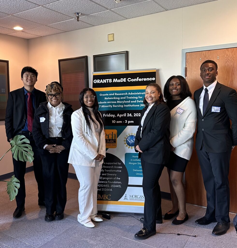 A group of six college students standing in front of a large posture board that says GRANTS MaDE Conference, with the description. The students have hand-written name tag stickers on their blazers. 