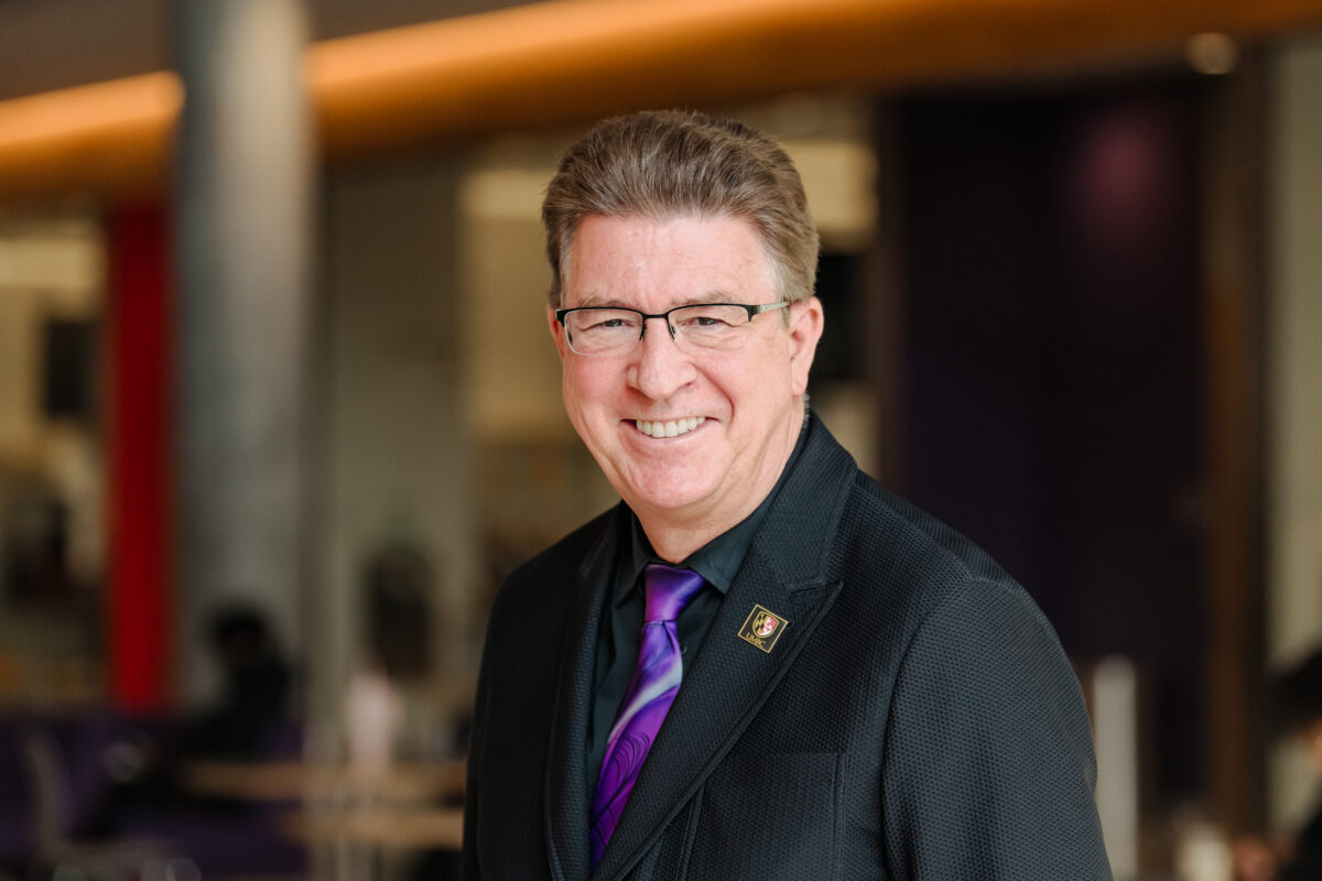 A man, Karl Steiner, wearing glasses smiling at the camera. He has a on a tie with purple swirl like design and a UMBC lapel pin clipped to his lapel. 