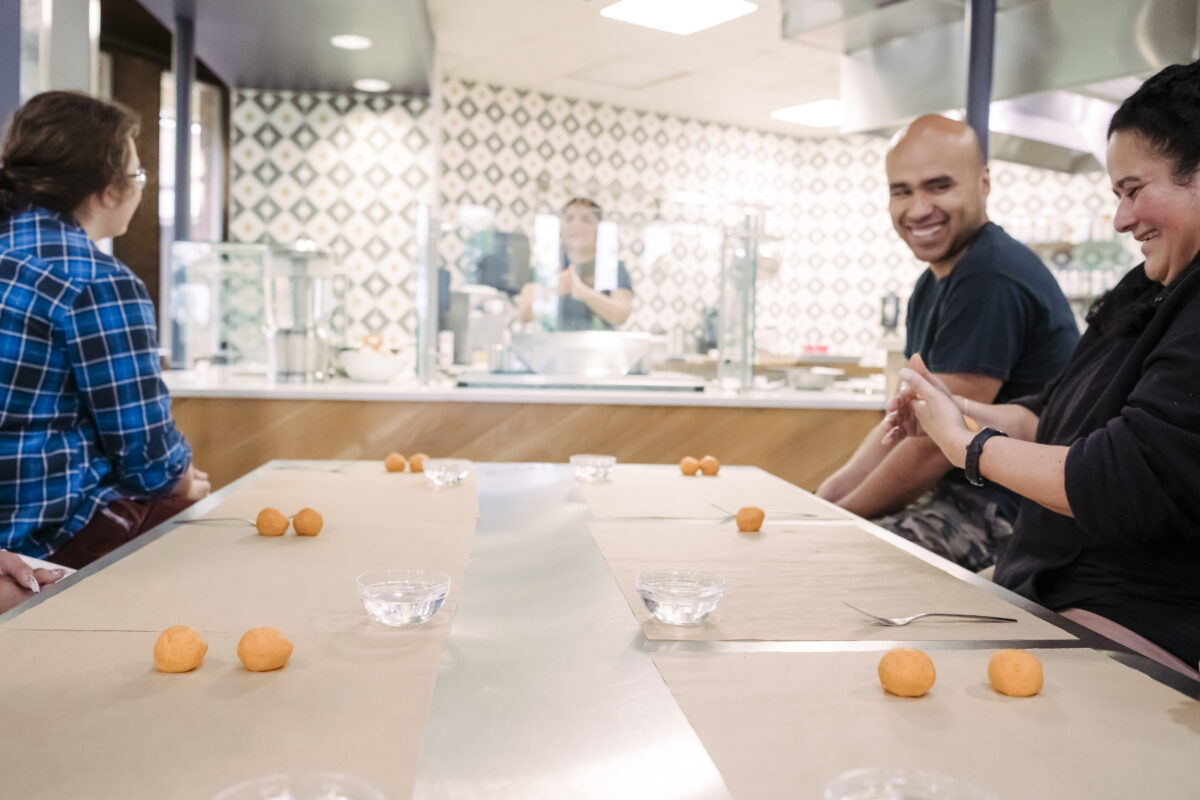 Three UMBC college students sit at a stainless steel table covered with butcher paper with small bowls of water, forks, and a mound of yellow dough with a SalviSoul chef Karla T. Vasquez cooking in the background at the True Grit's Test Kitchen.