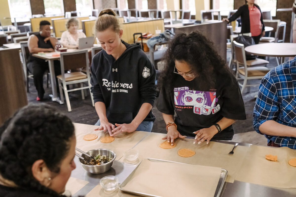 Students sit at a stainless steel table in a cafeteria shaping yellow dough into palm-sized circles