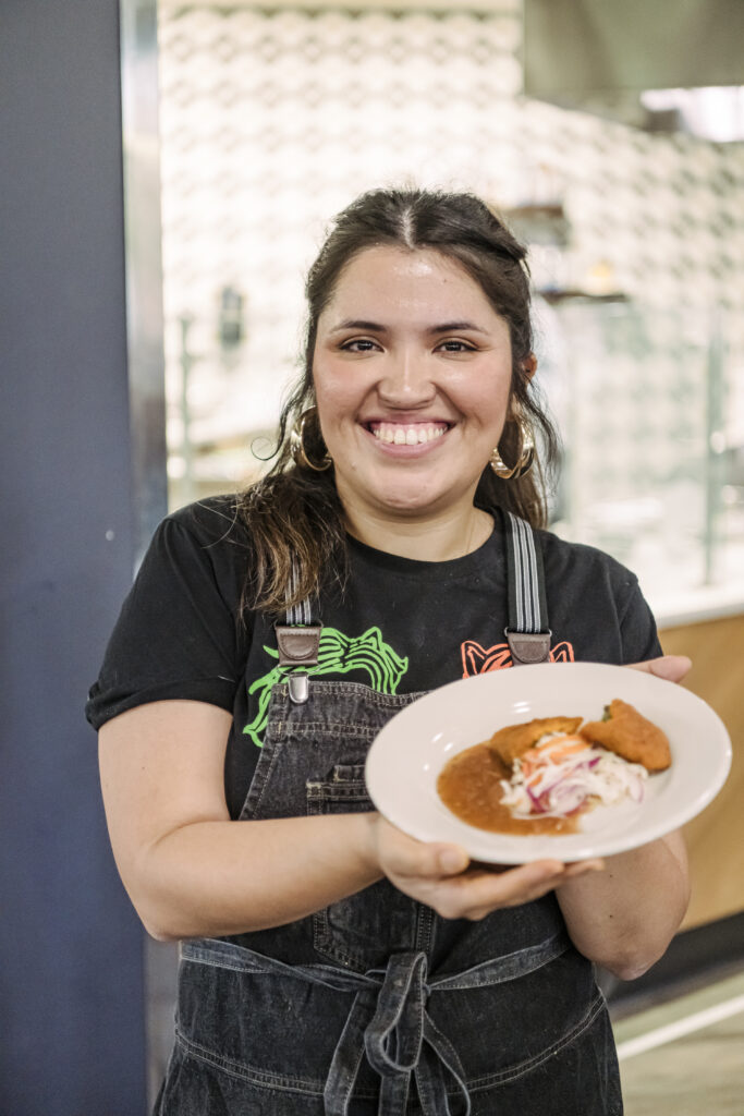 Karla T. Vasquez, a Salvadoran chef, stands in a cafeteria holding a white ceramic plate filled with a fried meat pie with slaw and salsa