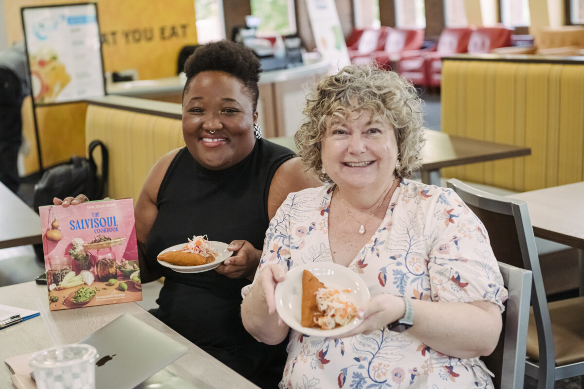 Two people sit next to each other holding up small plates of Salvadoran pastelitos, a small fried vegetable pie, at a cafeteria and holding the SalviSoul Cookbook by Karla T. Vasquez