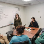 A professor sits with students to discuss research projects