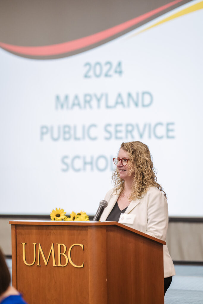 A woman stands at UMBC podium. Powerpoint in background reads "2024 Maryland Public Service Scholars"