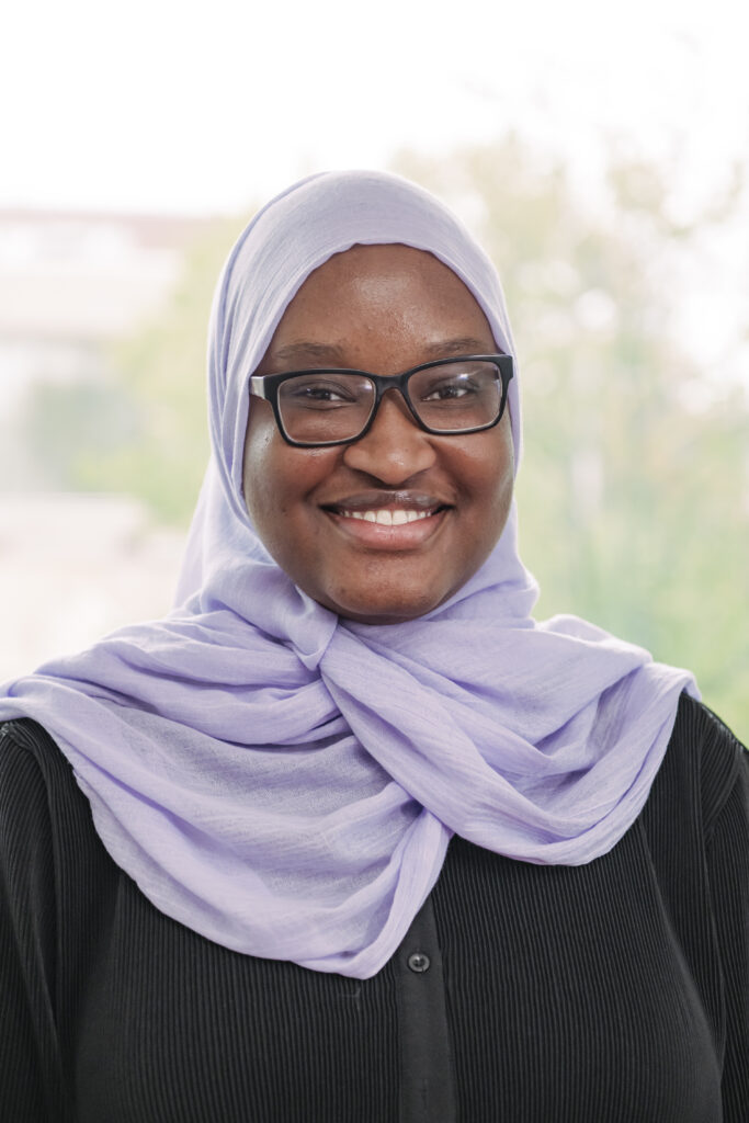 headshot of a smiling woman wearing a lavender headscarf and black glasses