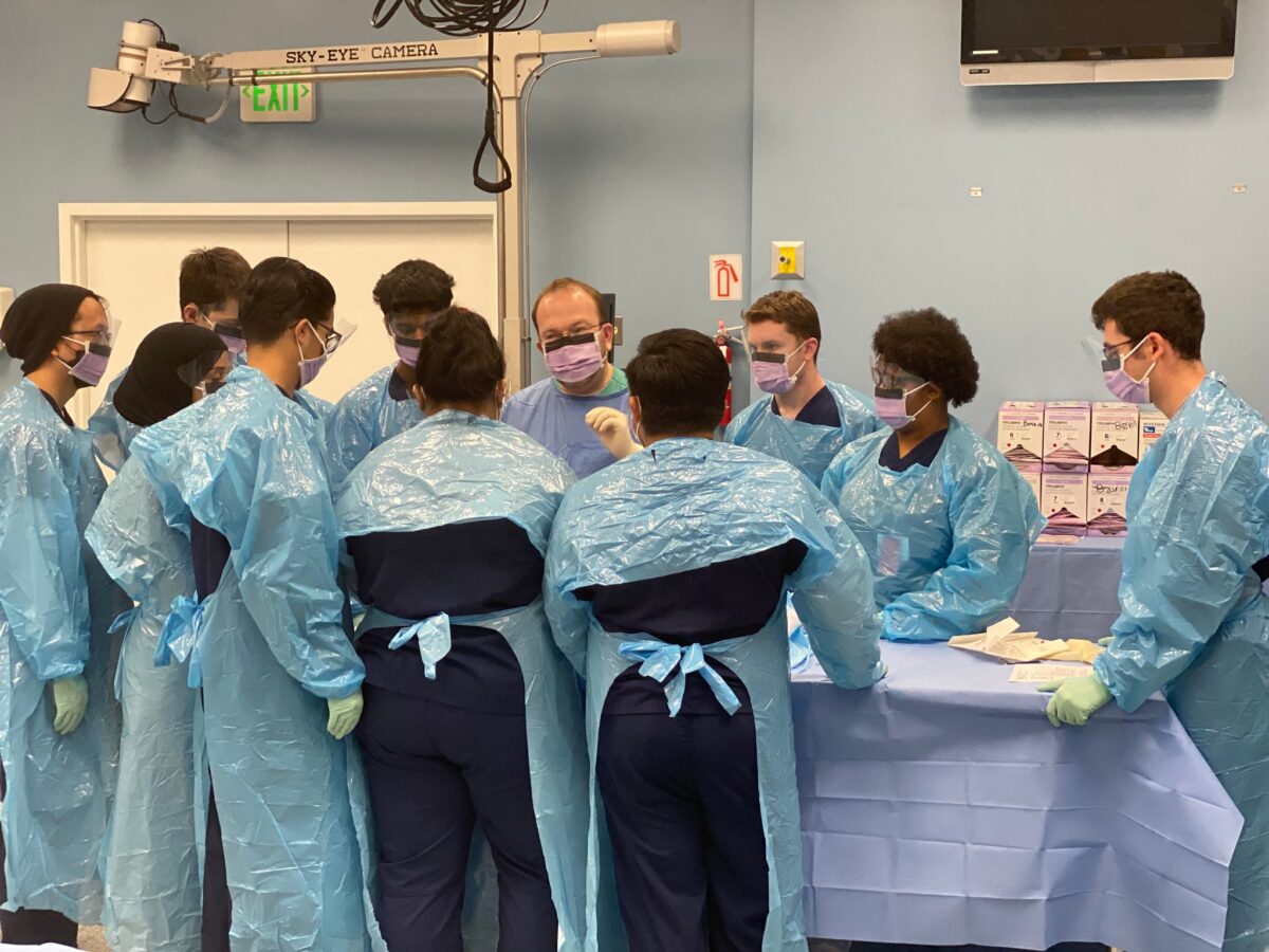 Matthew J. Levy with a group of medical students in scrubs stand around a hospital table teaching about emergency disaster and health systems