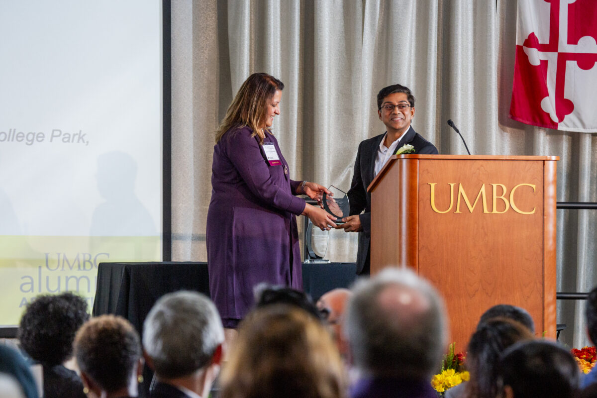 a man received an alumni award from a woman on stage behind a podium that says UMBC