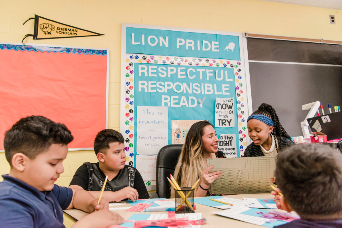 An elementary school student reviews a math chart with the teacher who is sitting at a table with other students MSDE 