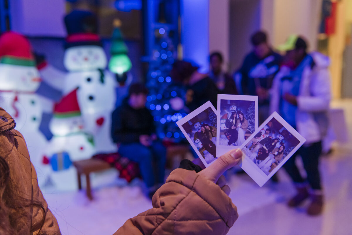 Close-up of a hand holding three polaroid pictures taken at (seb)'s Winter Wonderland event. In the background, there festive decorations like inflated snowmen and a lit Christmas tree. in front of the decorations is a bench where people are sitting and appear to be bundled up for the cold weather