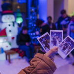 Close-up of a hand holding three polaroid pictures taken at (seb)'s Winter Wonderland event. In the background, there festive decorations like inflated snowmen and a lit Christmas tree. in front of the decorations is a bench where people are sitting and appear to be bundled up for the cold weather