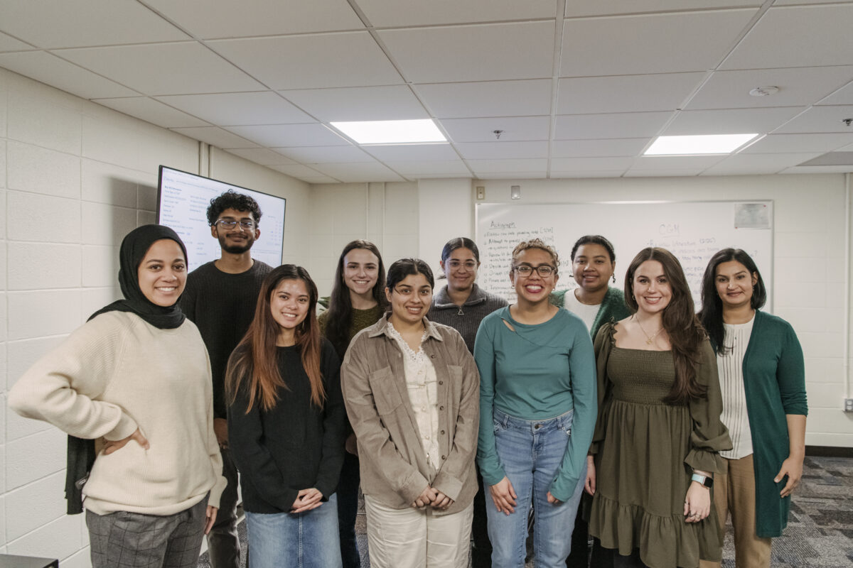 A group of eight college students and a professor stand together fin a classroom for a group photo in a classroom