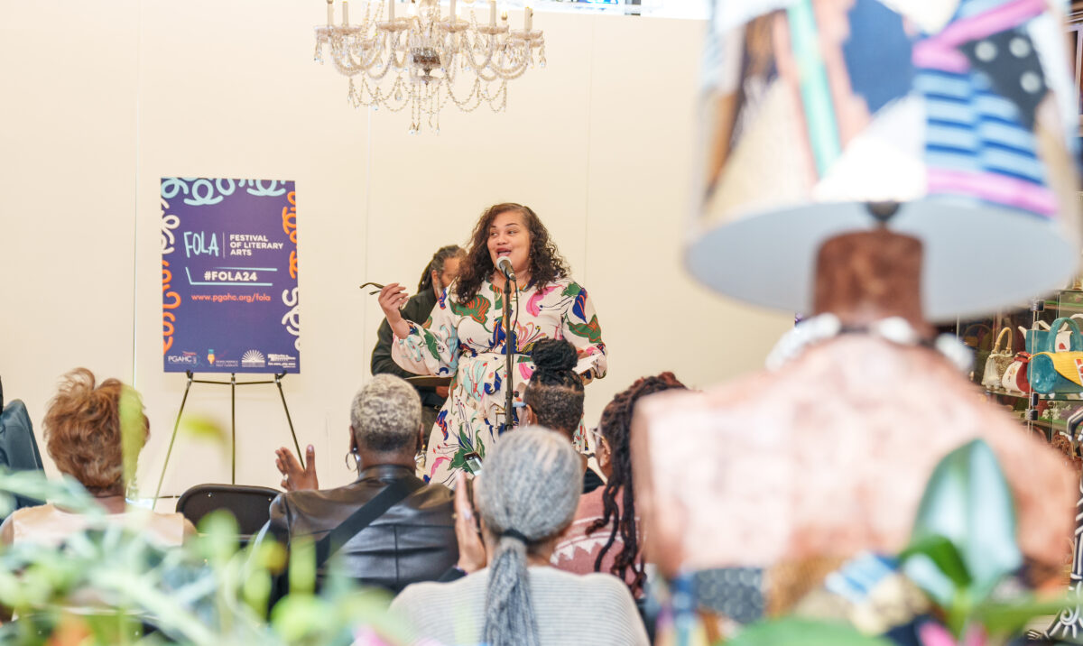 A woman stands at a microphone using her voice to give a presentation to a room of people. There is a sign propped up that reads "Festival of Literary Arts"