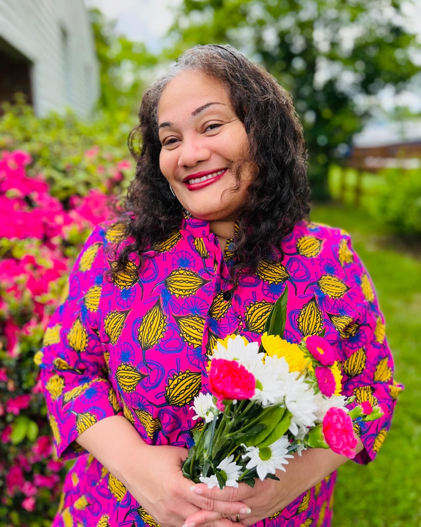 A woman smiling wearing a brightly patterned pink dress, holding a bouquet of flowers, while standing in front of flowers.