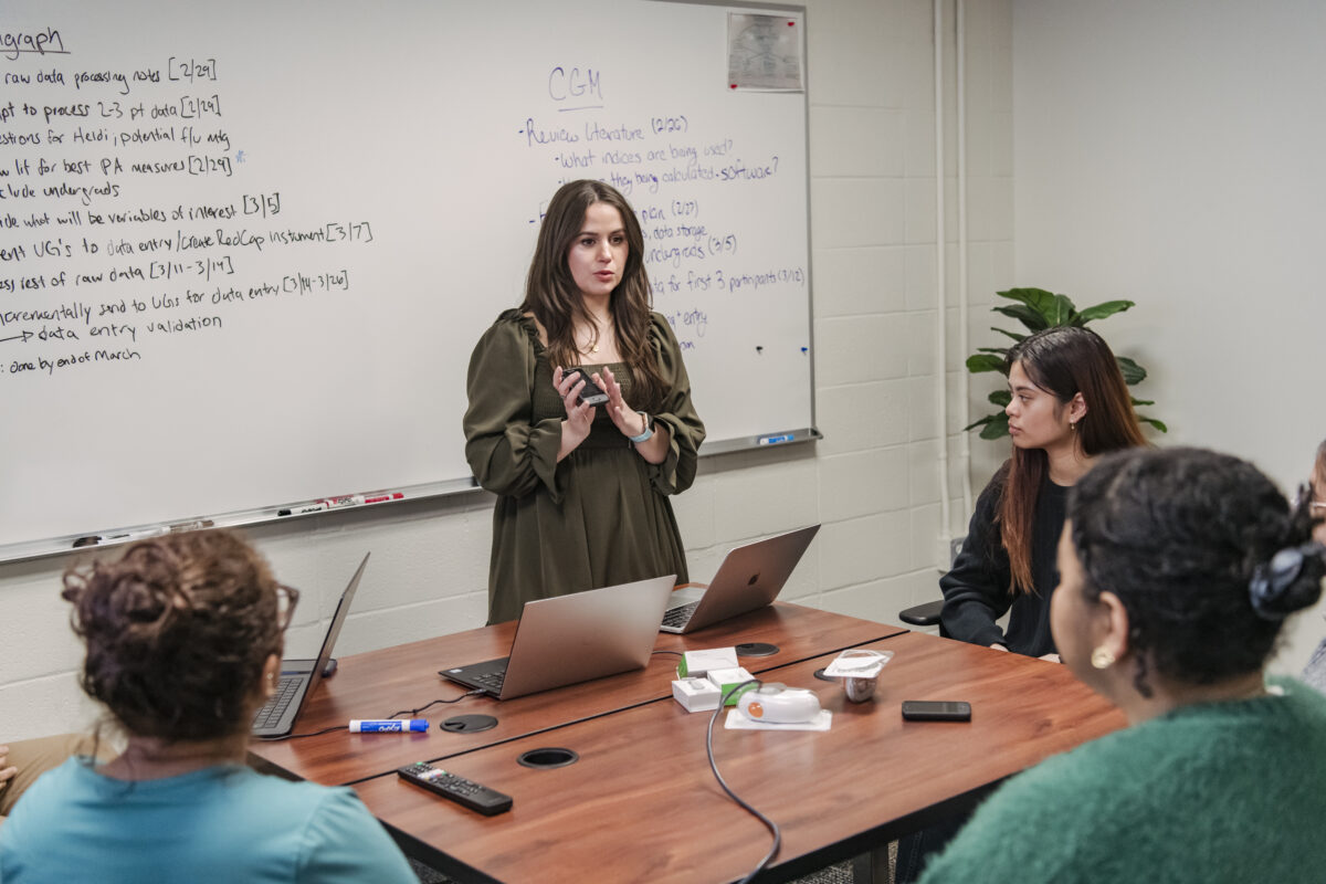 A graduate students stands in front of a group of students explaining a small glucose monitor works