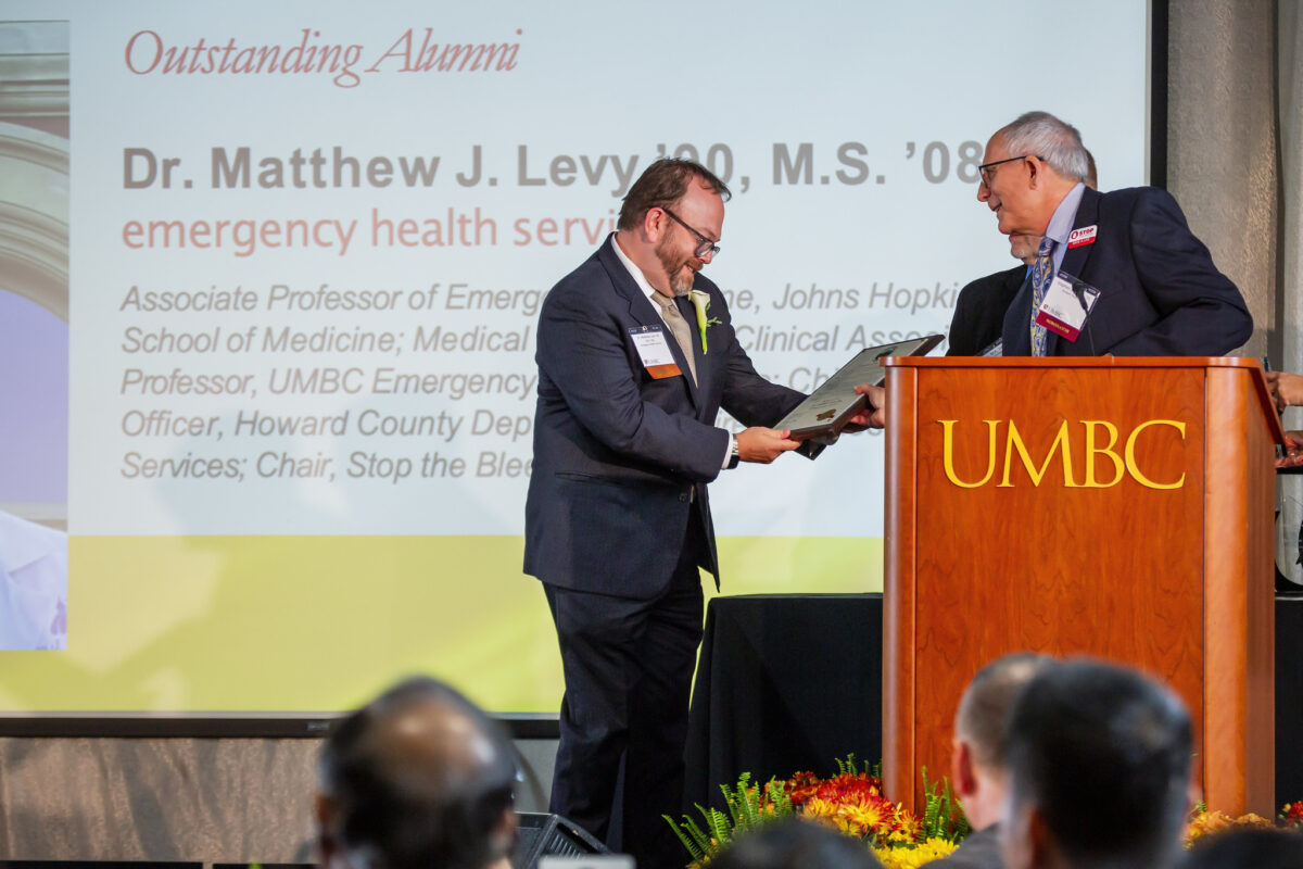 Matthew Levy person on stage receives an award from a person at a podium with a description of the award on a projection screen in the background