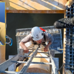 view from above - person climbing up ladder wearing construction helmet; two feet wearing boots visible on an upper platform above the climber's head; inset shows full height of tower being climbed, view up from the base