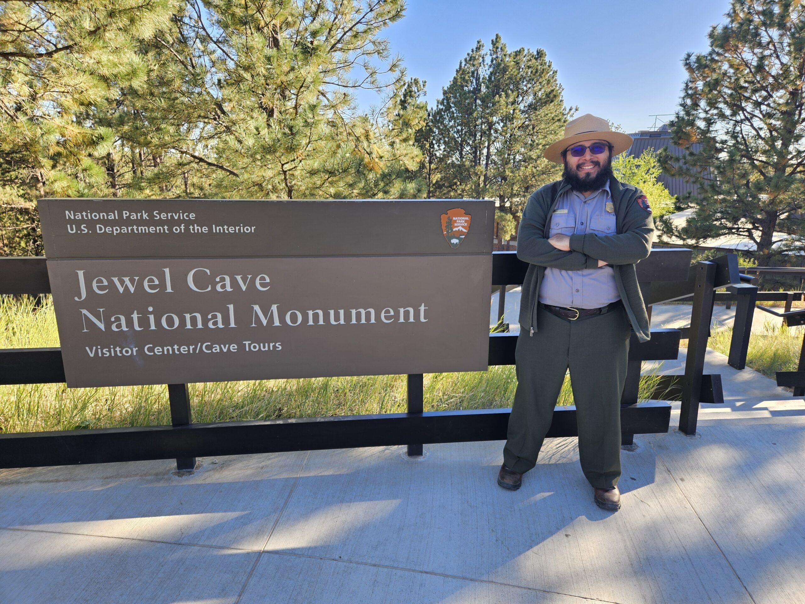 Jeff Garcia, a park ranger, stands with arms crossed in front of the Jewel Cave National Monument sign. The background features pine trees and a sunny outdoor environment