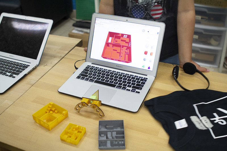 A computer screen display a design file. 3D printed objects are arrayed in front of the computer.