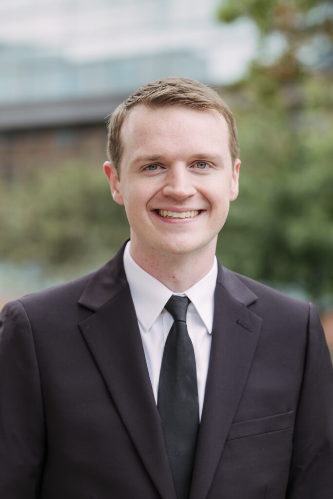 Collin Sullivan ’19, wearing a dark suit and black tie, smiles confidently in a professional outdoor portrait. 