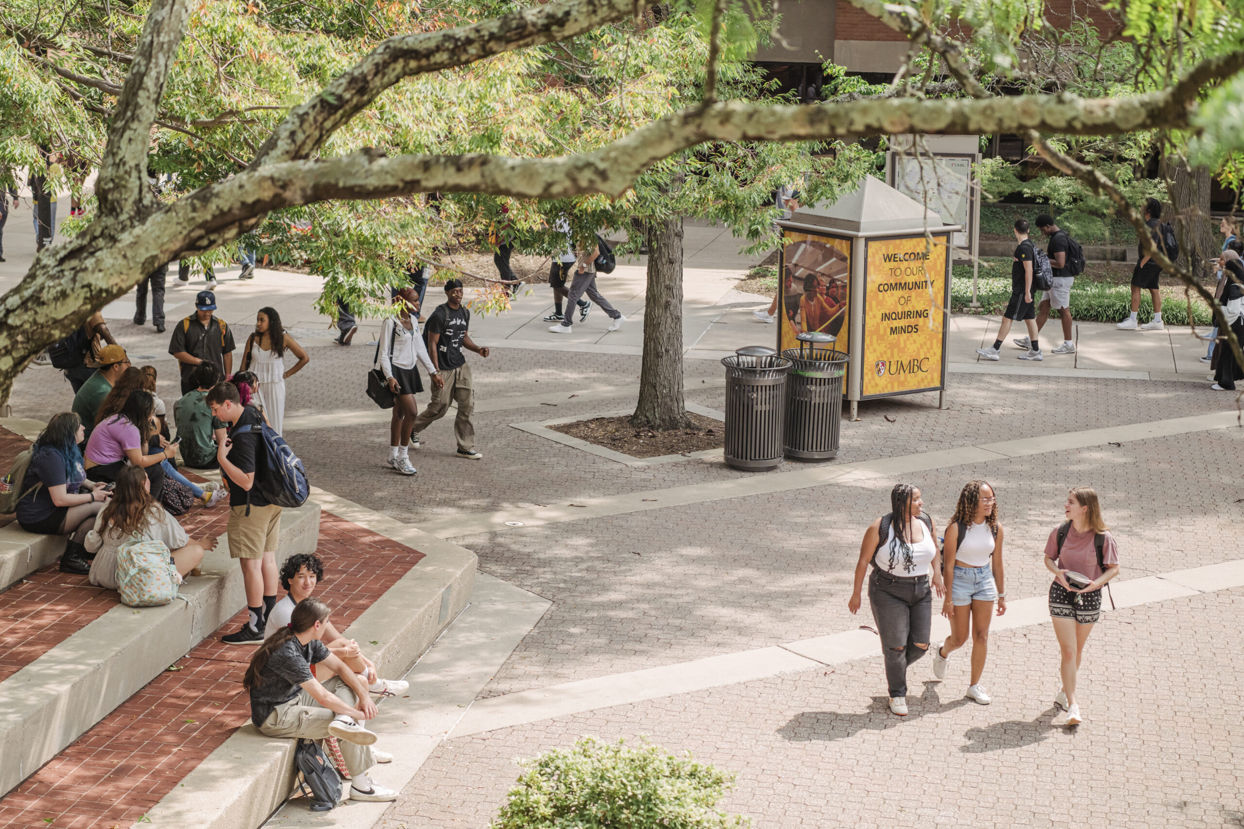 UMBC students walking through Academic Row, some students are sitting on steps on the left side of the image.