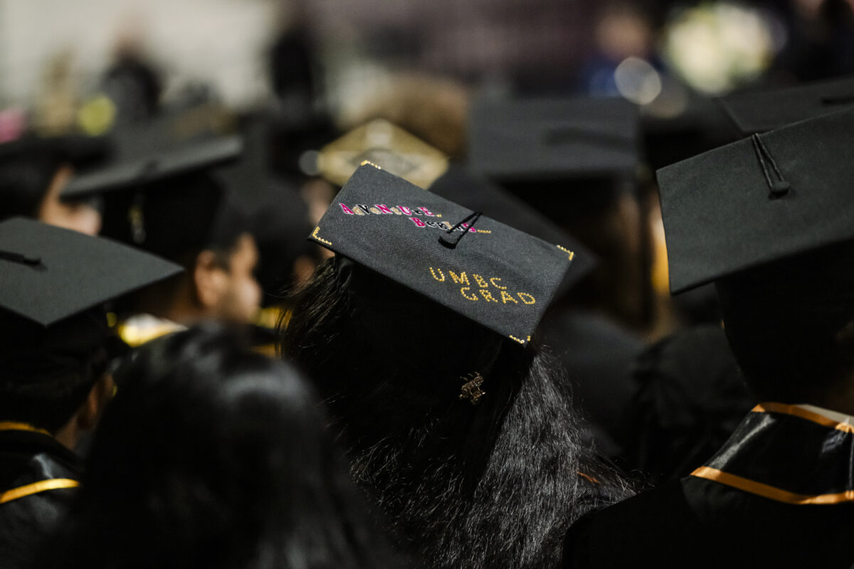 Graduates in black caps and gowns, with one cap featuring "UMBC Grad" text, seated in an auditorium.