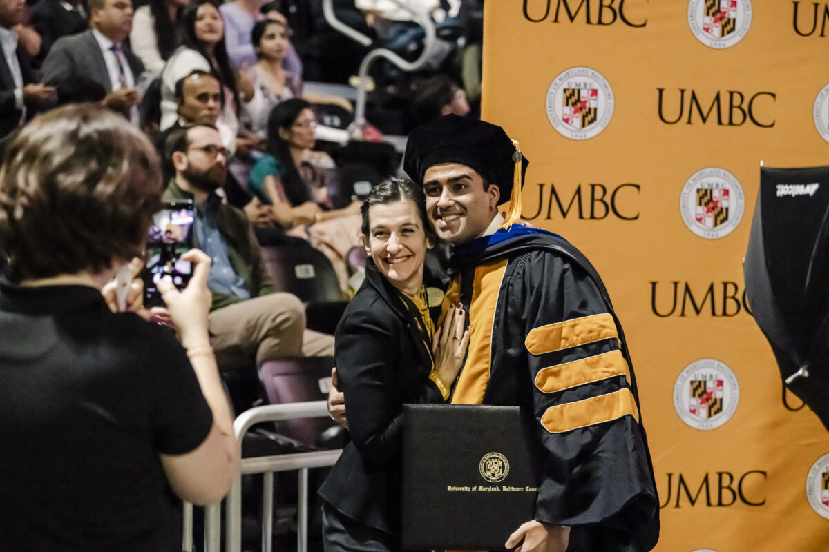 A graduate in cap and gown poses with a woman for a photo during a UMBC ceremony. Others are seated in the background.