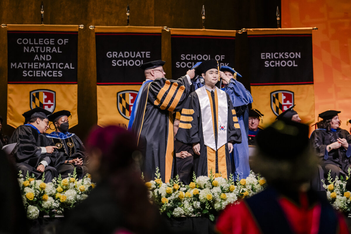 A graduate in a black cap and gown is being hooded on stage during a ceremony. The stage is decorated with banners from various schools, and flowers are arranged at the front. Other officials in academic regalia are seated and watching.