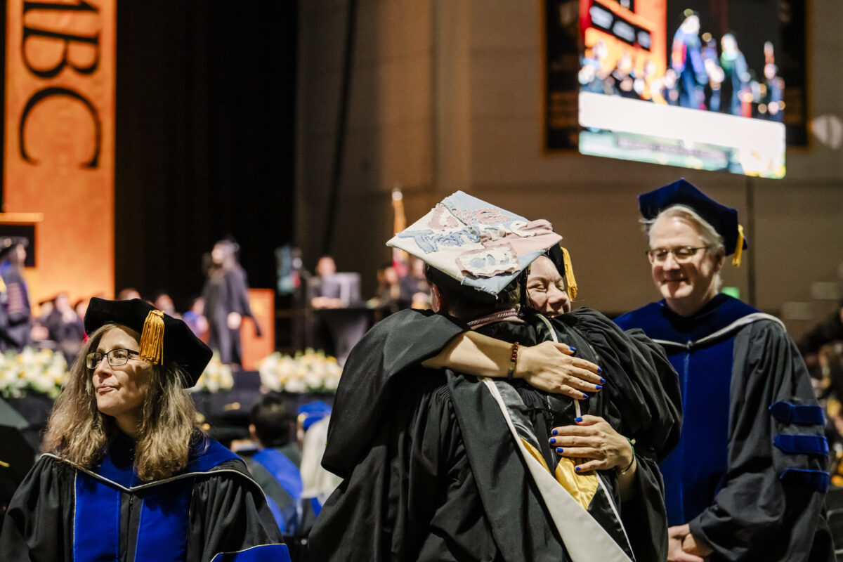 Graduates in caps and gowns shake hands with faculty in academic regalia at a graduation ceremony. A crowd of spectators is seated in the background.