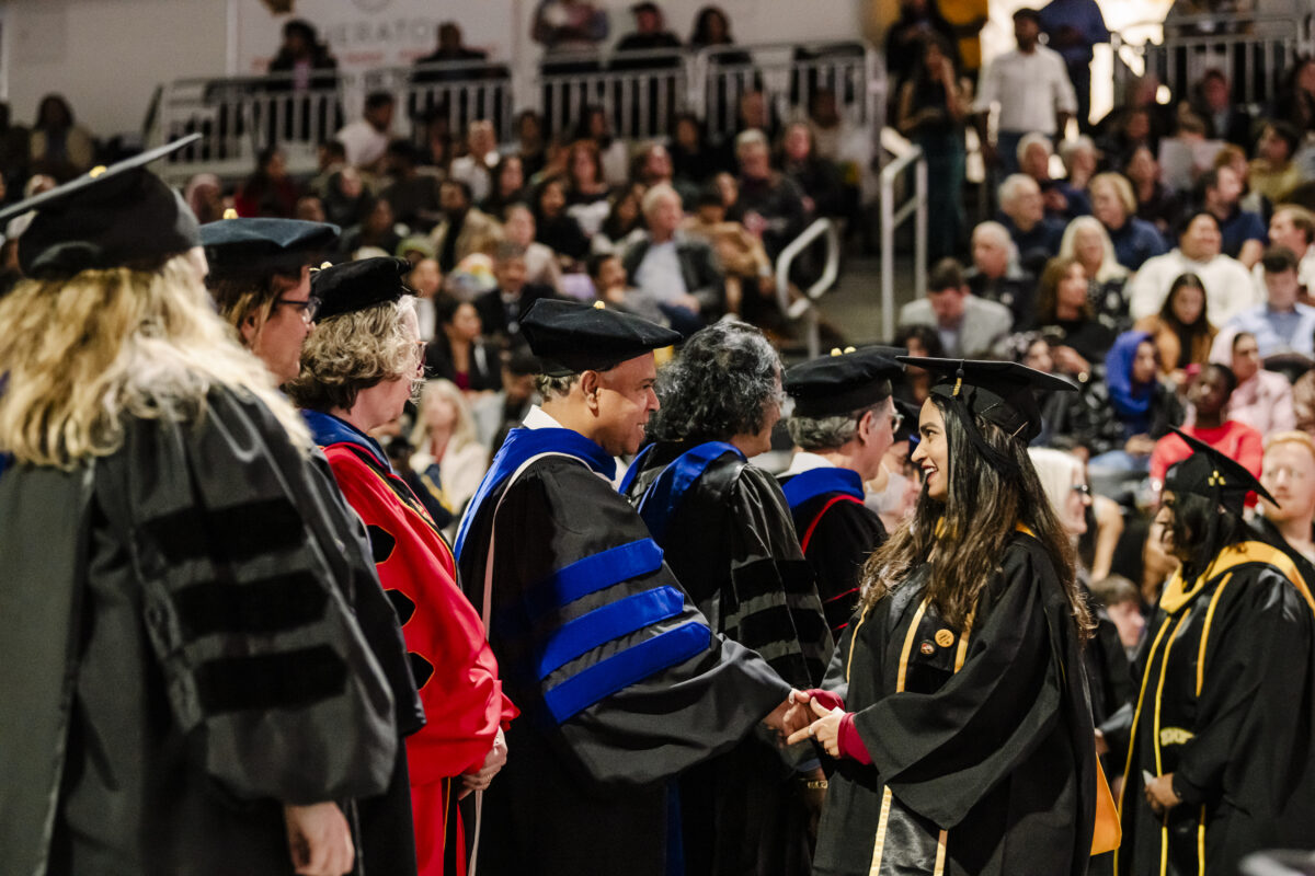 Graduates in caps and gowns shake hands with faculty in academic regalia at a graduation ceremony. A crowd of spectators is seated in the background.