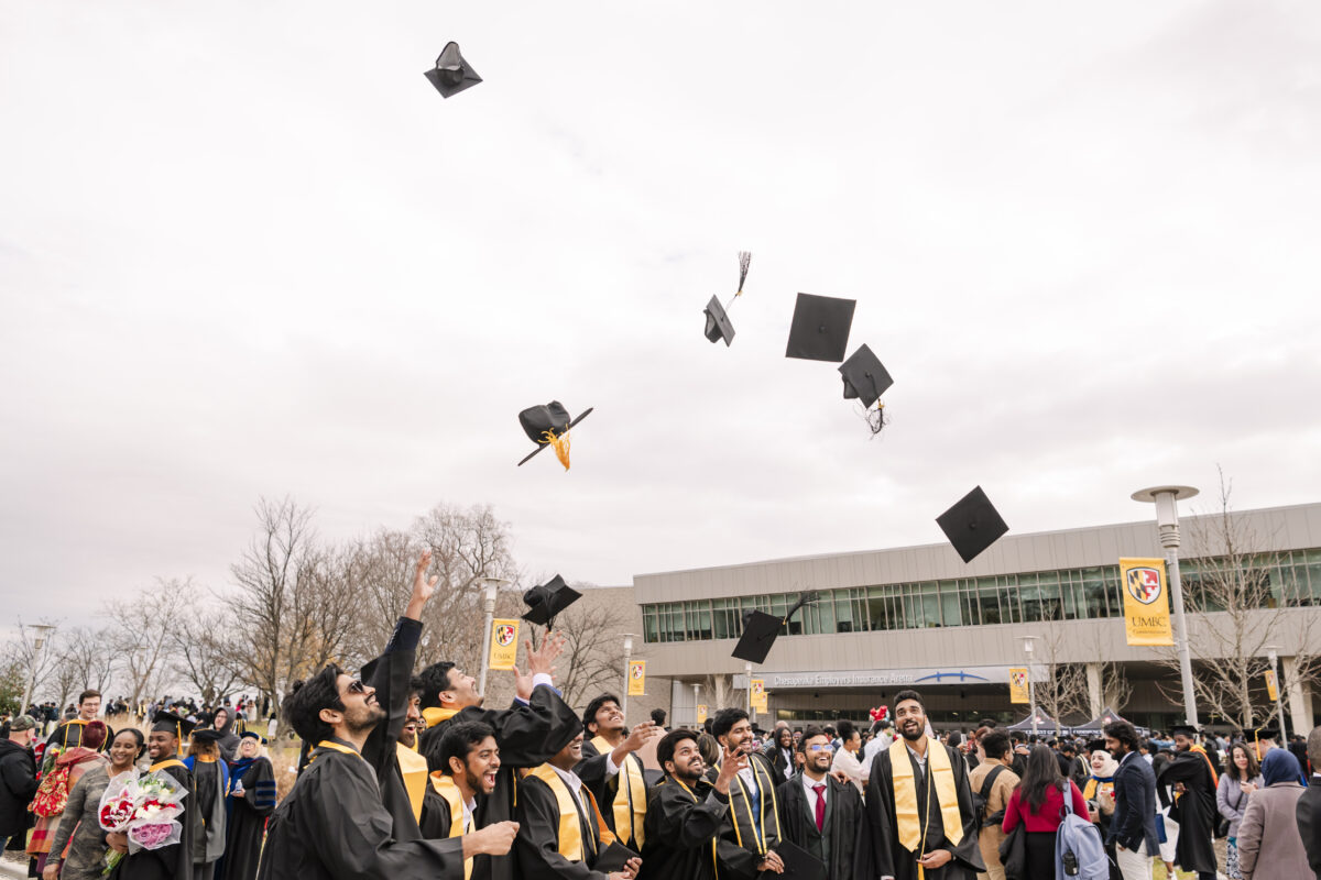 New graduates in caps and gowns celebrate by throwing their caps into the air outside a modern building. The sky is overcast, and people are gathered around, some taking photos. Banners line the path in the background.