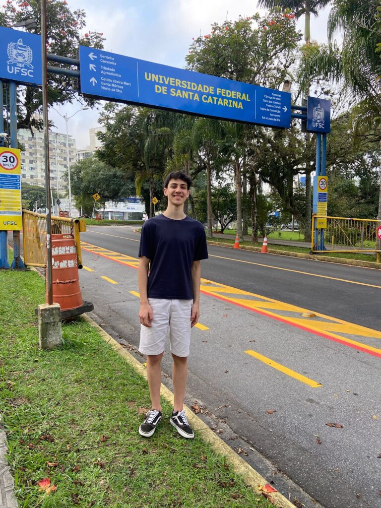 A Brazilian college student standing in front of a blue and gold directional sign with the name of a university global