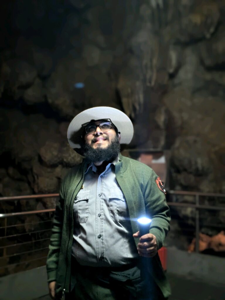 Jeff Garcia, wearing a park ranger uniform and hat, holds a flashlight while smiling inside Jewel Cave. The cave walls surround him as he stands underground.