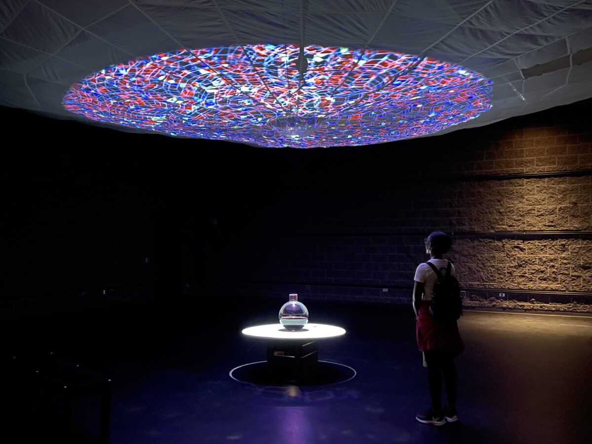 A circular bottle sit on a podium. Colorful light are projected on the ceiling. A person with a backpack looks at the display.