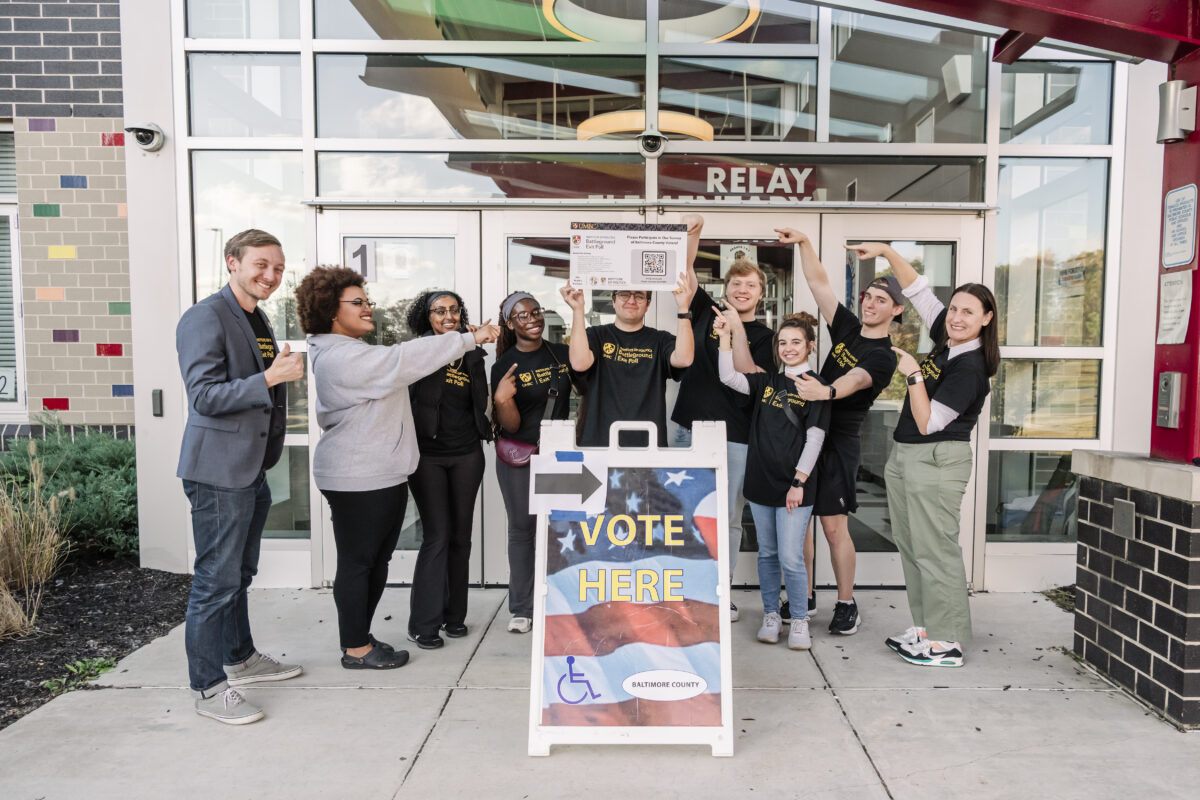 A group of seven college students wearing black and gold shirts stand with two faculty members in front of a public voting location behind a vote here sign. Battleground Exit Poll