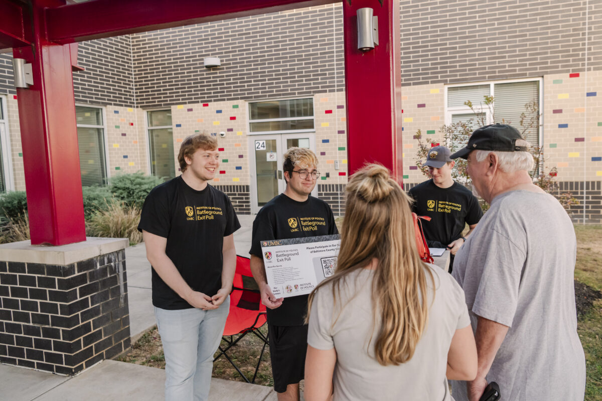 Three college students stand outside a voting station sharing a sign about a survey with voters.