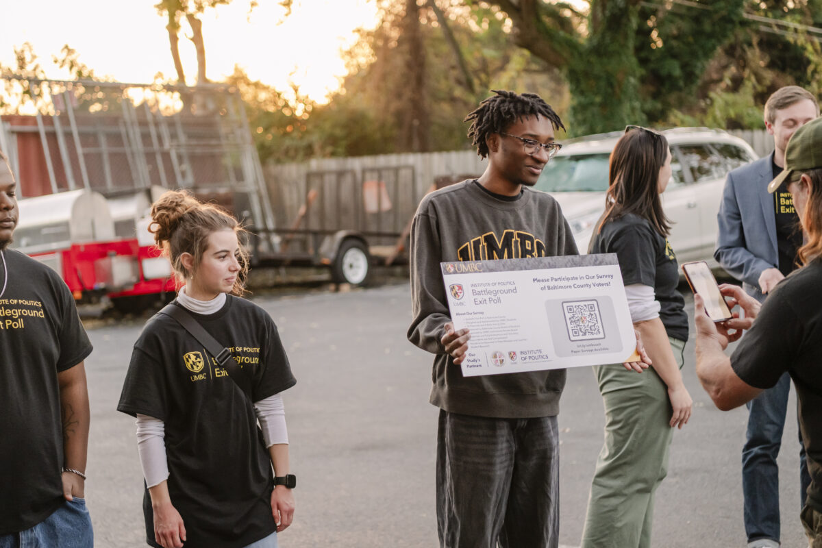 A college student wearing a grey UMBC sweatshirt stands outside a voting station holding a sign with a QR code for the Battleground Exit Poll