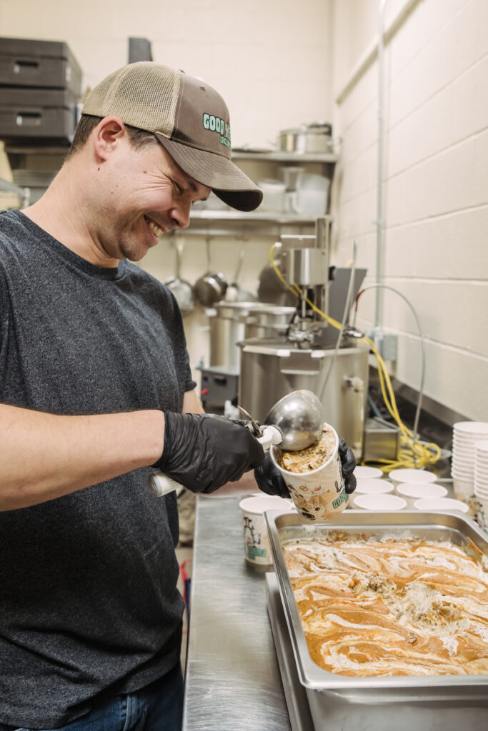 a man in a black shirt smiles while he scoops ice cream from a large tray into a pint container