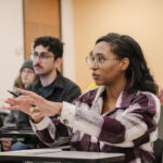 A college student in class sits at a desk speaking with the professor.