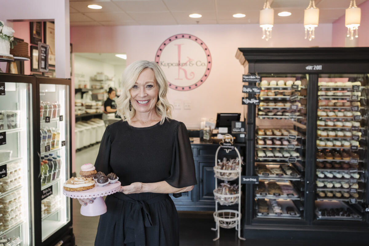 a woman in a black dress shirt stands in a pink bakery surrounded by sweets holding a tray of cupcakes