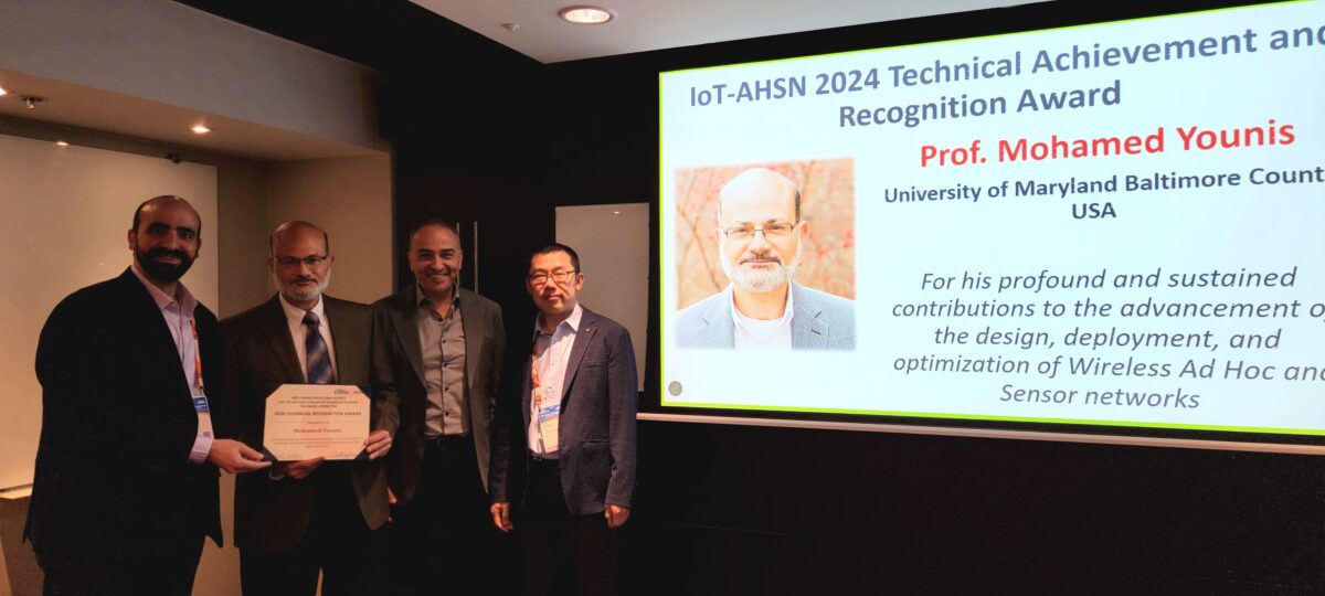 Four men stand in group. One presents a certificate to another. Screen displays slide that reads "IOT-AHSN 2024 Technical Achievement and Recognition Award"