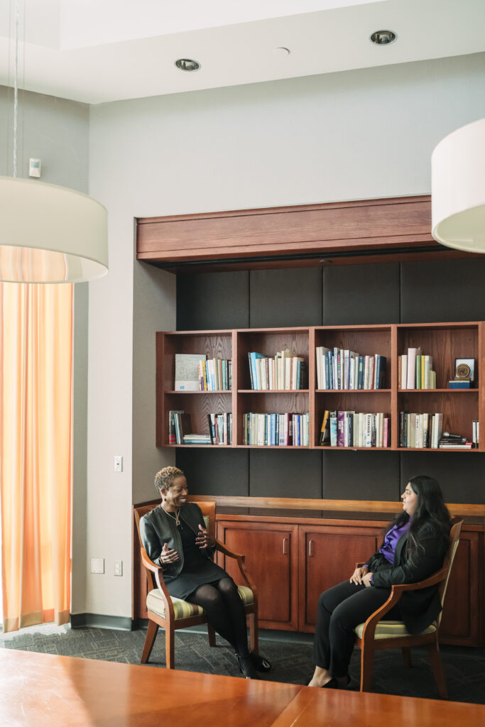 two women sit next to a bookshelf in nice wooden chairs during office hours