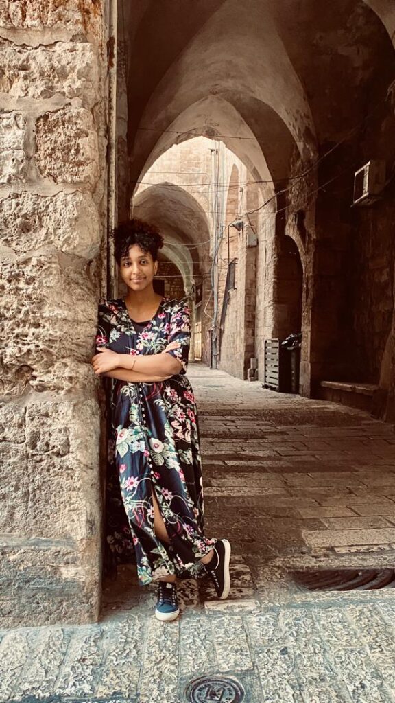 Nardos Amanuel Kebede with a floral dress stands in a stone breezeway with large stone arches in Jerusalem 