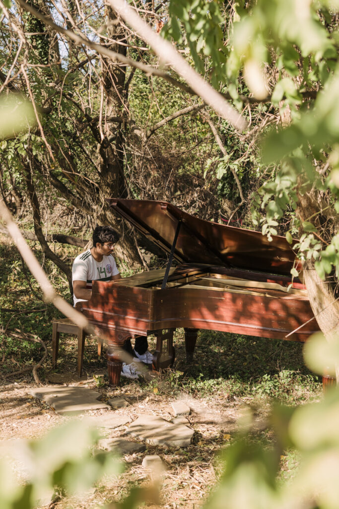 a young man in a white jersey plays at a piano tucked away in the woods