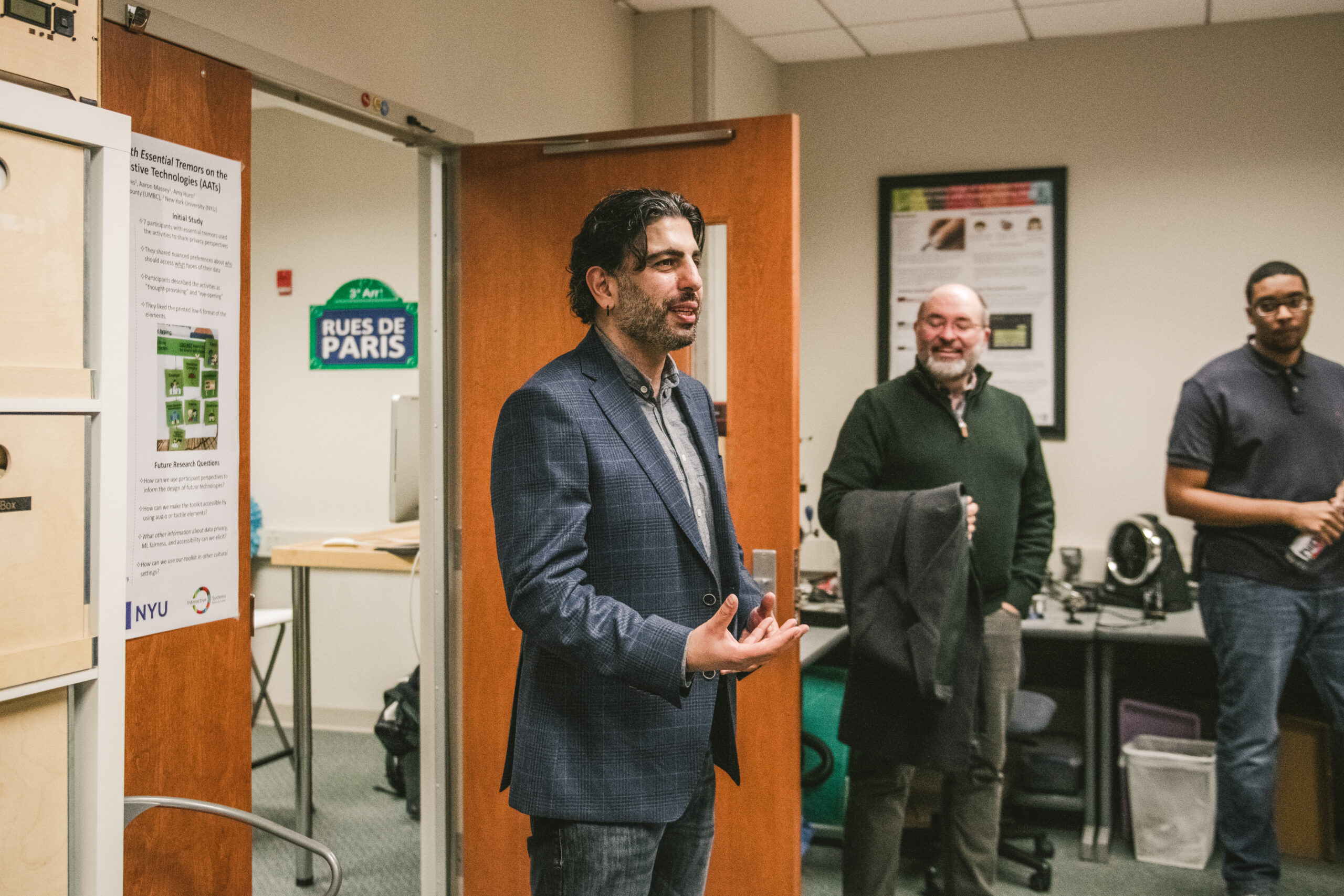 Man in button-up shirt and suit coat speaks with other people in front of an open lab door.