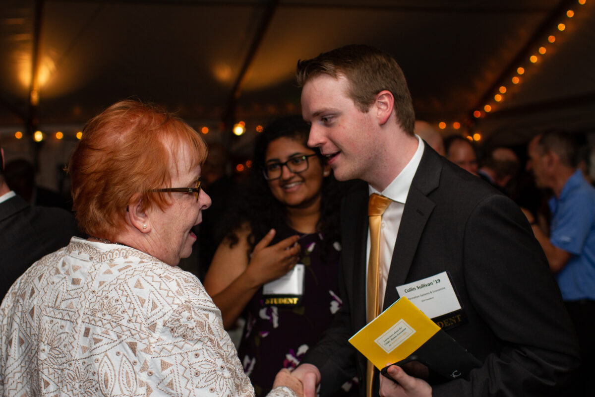 Collin Sullivan ’19, Digital Credential leader, wearing a black suit and gold tie, shakes hands with an older woman in a white patterned jacket at an event. The interaction is warm and professional, with Collin holding a yellow program booklet. In the background, a woman with curly hair and glasses smiles, adding to the lively atmosphere of the gathering