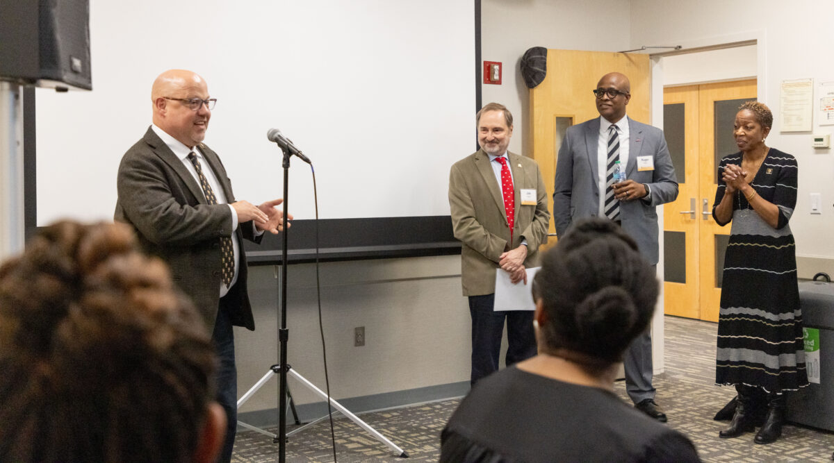 man stands speaking at a microphone, three people stand off to the side, others are seated listening.