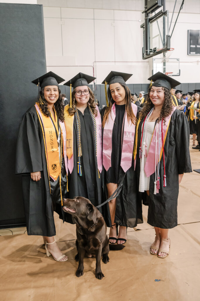 Four graduates in caps and gowns stand together with a brown dog at a graduation ceremony. They wear various honor cords and stoles, and are smiling inside a large hall with other graduates in the background.