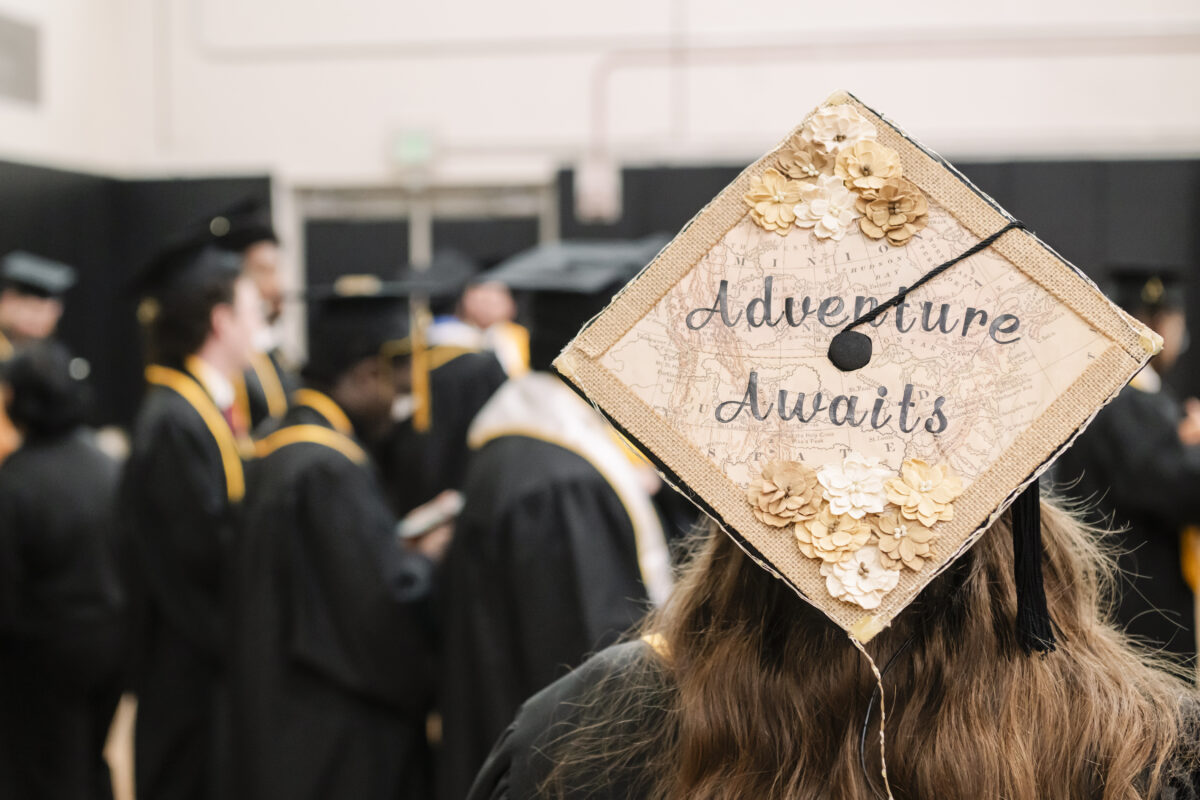 A graduate wearing a decorated mortarboard with the words Adventure Awaits, adorned with paper flowers and a map design. Other graduates in black robes and caps are gathered in the background, preparing for the ceremony.